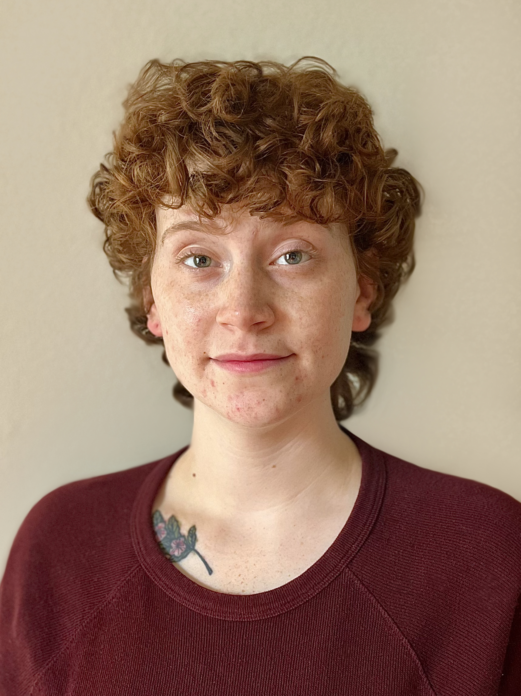 A headshot of Laurel Greenway. A white female with freckles and short curly ginger hair, in a dark maroon sweater that is slightly covering a tattoo of a laurel branch on her right collar bone.