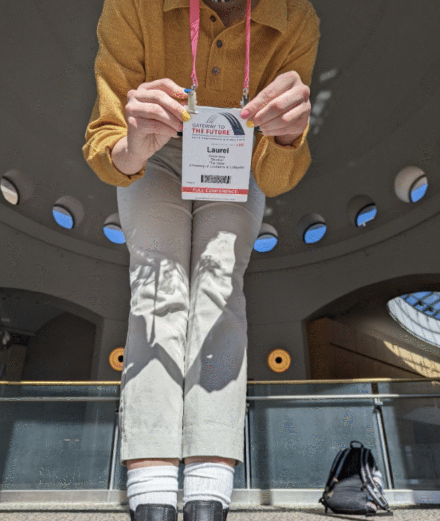 A photo of Laurel in a yellow sweater and white pants showing off her tag for The United States Institute for Theatre Technology.